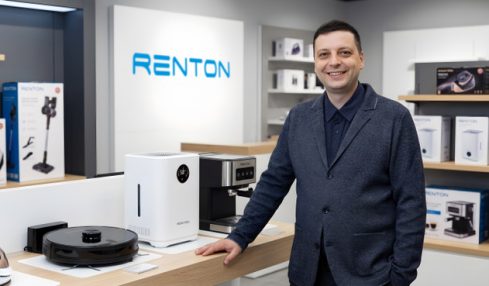 Man in a suit standing at a display counter with electronic devices in a store, Renton sign in background.
