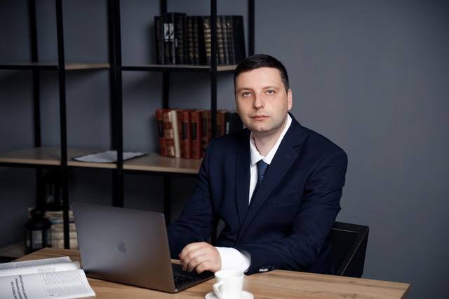 Man in suit sitting at desk with laptop and books in background, gray wall, professional setting.