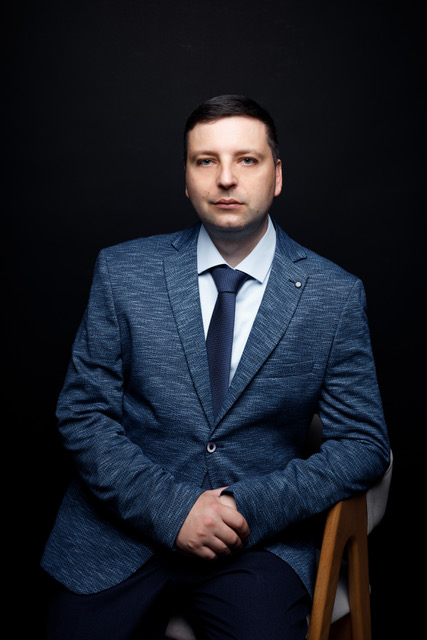Man in a blue suit and tie sitting on a chair against a dark background.