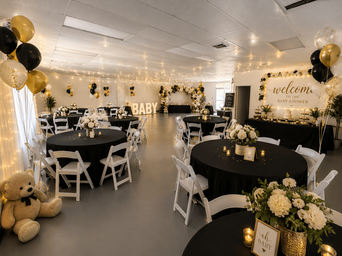 Decorated event room with black and gold balloons, floral arrangements, and a welcome sign, set for a celebration.