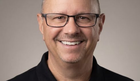 Smiling man with glasses and short hair, wearing a black collared shirt, against a plain background.