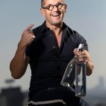 Man with glasses smiling, pointing to his head, holding a water bottle, sitting on a stool against a plain background.