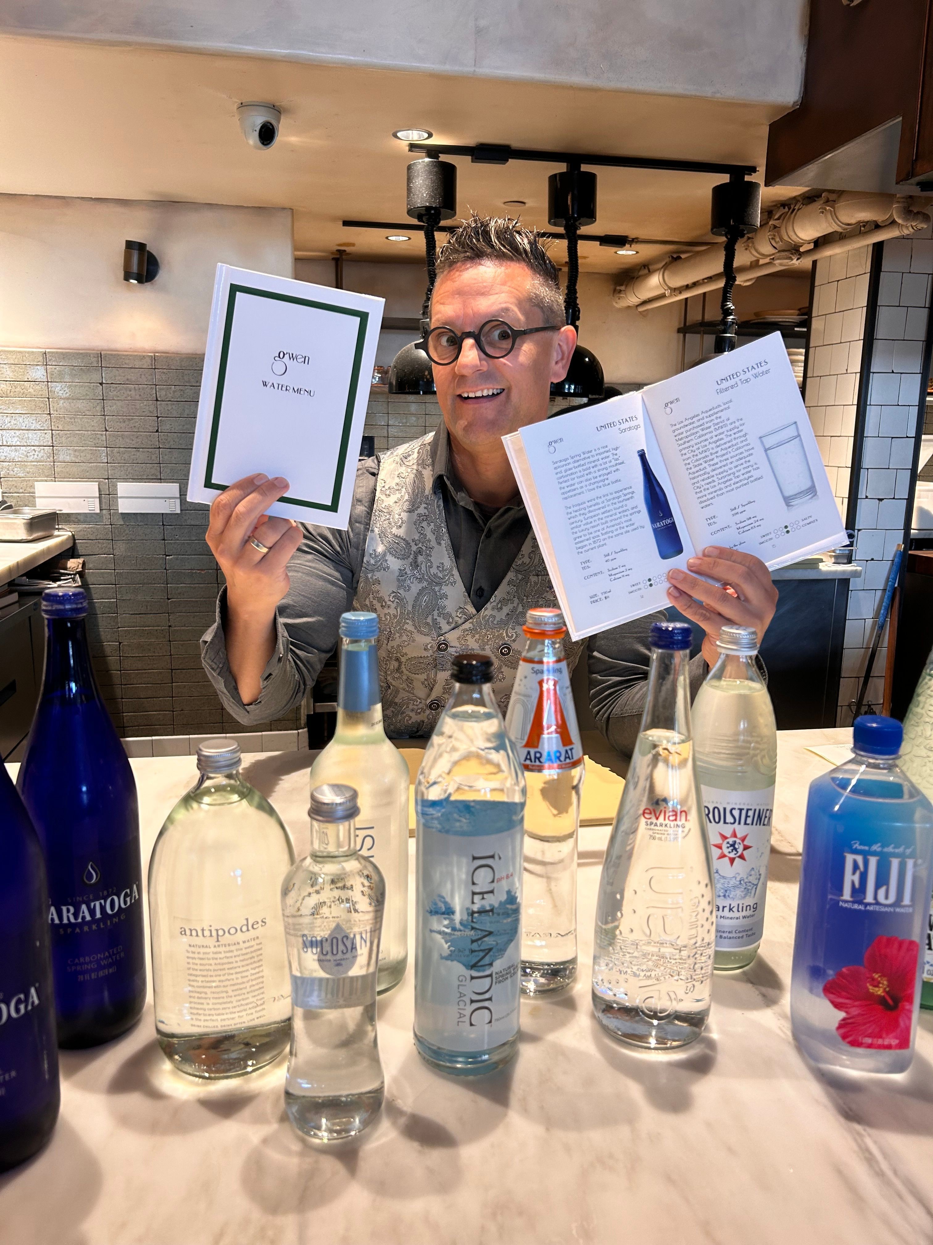 Person with glasses holding a book and a framed certificate, sitting at a table with bottles of alcohol, in a room with brick walls.