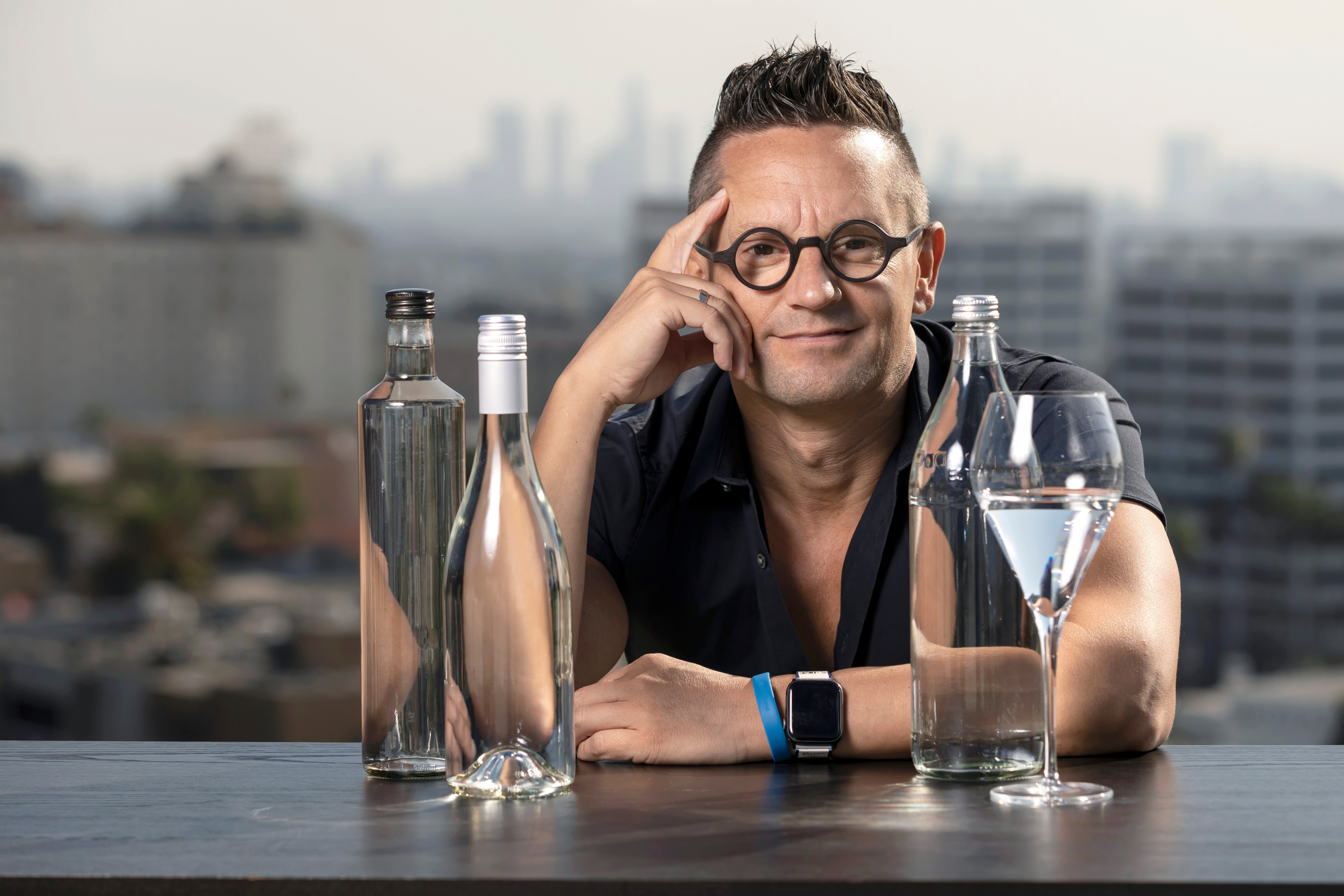 Person with glasses and short hair sitting at a table with three bottles and a glass, cityscape background.