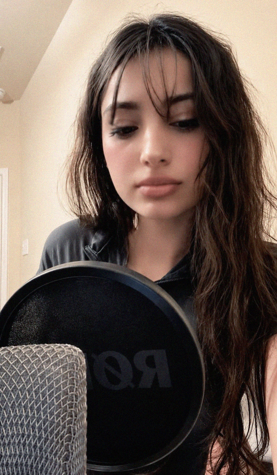 Young woman with long, wavy dark hair looking down, holding a microphone, indoors with beige walls.