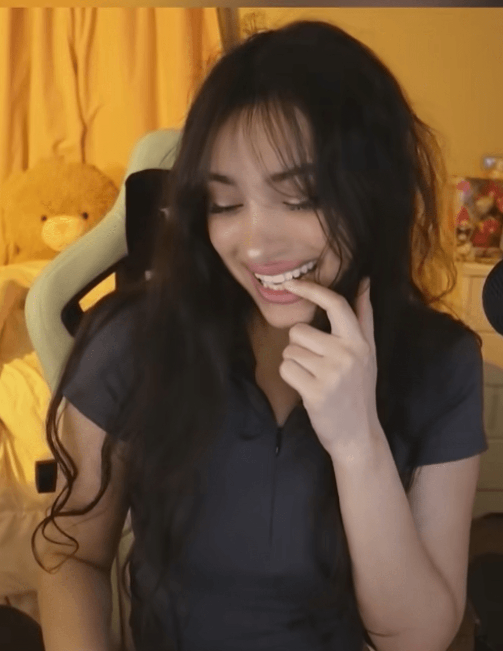 Young woman with long dark hair smiling and touching her chin, sitting indoors with a yellow curtain in the background.
