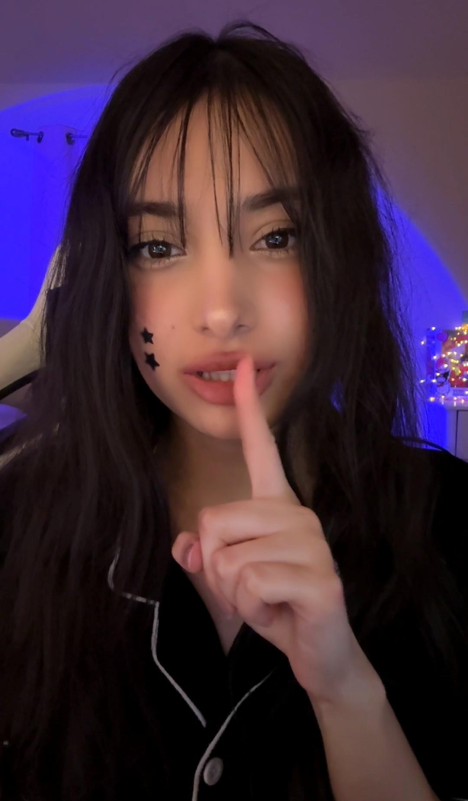 Young woman with long dark hair holding finger to lips, with small star-shaped face stickers, in a dimly lit room with blue lighting