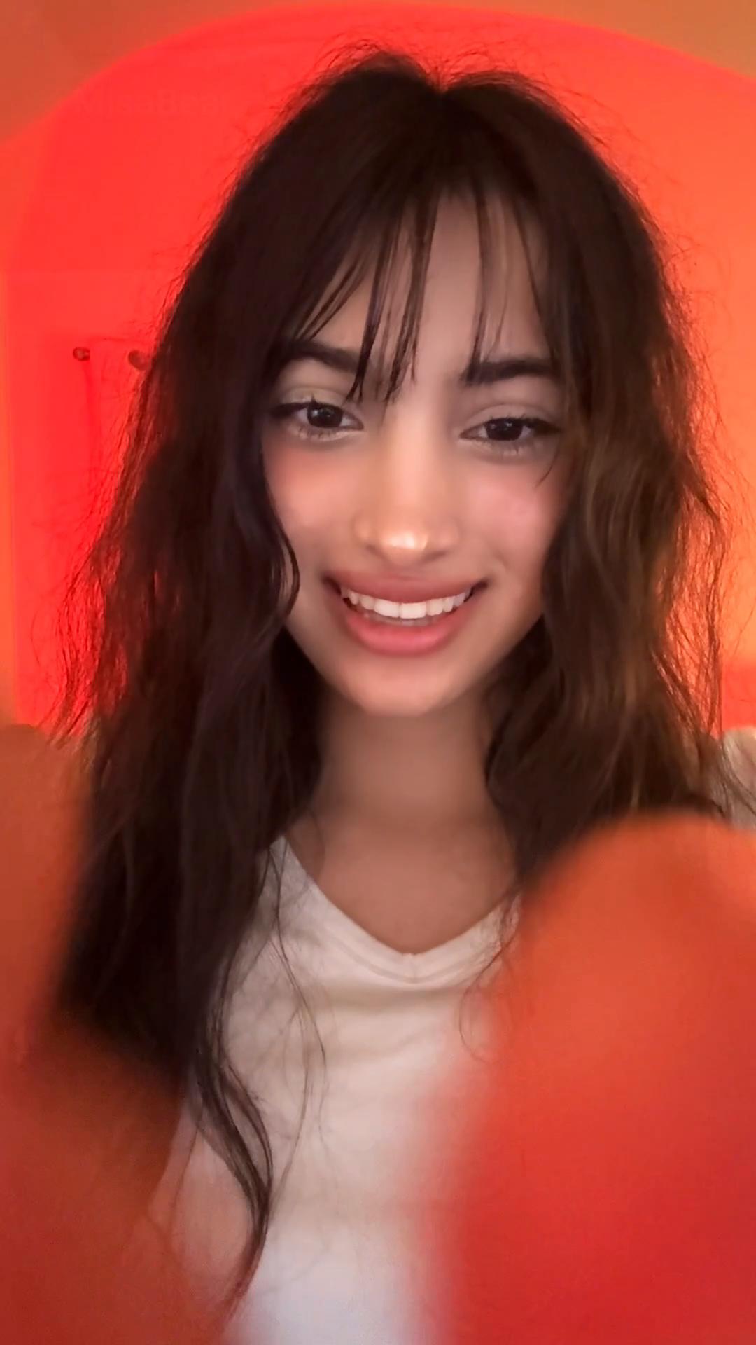 Young woman with long wavy hair and bangs smiling, wearing a white shirt, against a red background.