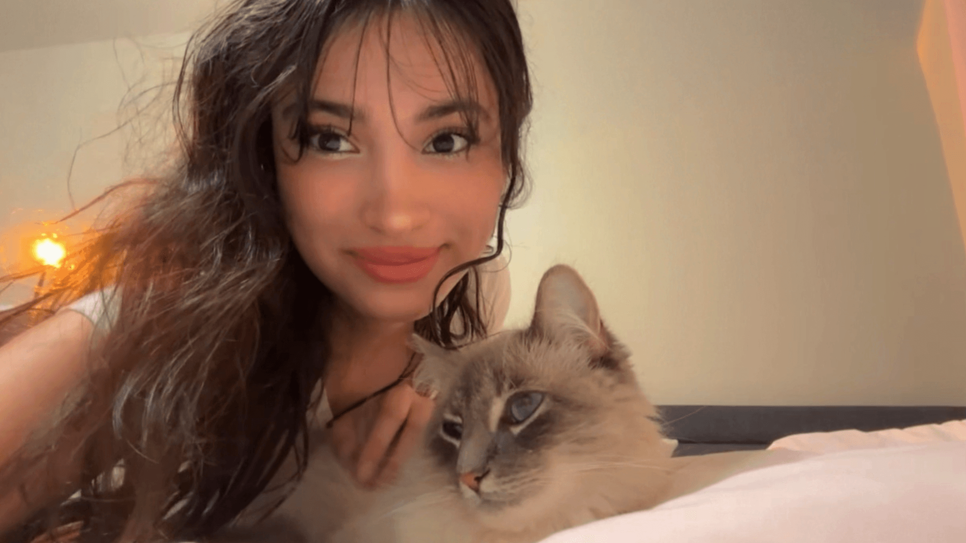 Young woman with long dark hair holding a light-colored cat, both lying down indoors.