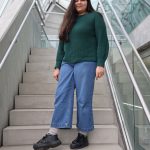 Woman with long hair wearing a green sweater and blue jeans standing on stairs inside a modern building.