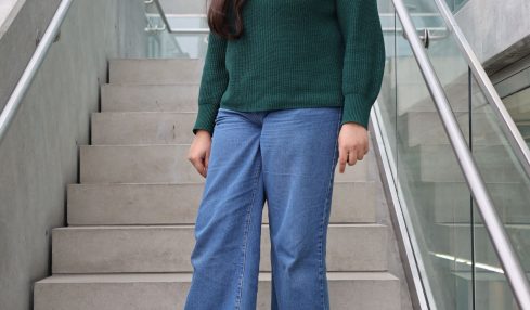 Woman with long hair wearing a green sweater and blue jeans standing on stairs inside a modern building.
