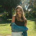 Young woman with long hair standing outdoors in sunlight, wearing a black top and denim jacket, smiling.