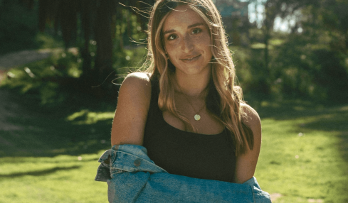 Young woman with long hair standing outdoors in sunlight, wearing a black top and denim jacket, smiling.