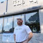 Man with a beard and white cap standing outside gym with glass windows and sign reading 'The Gym'.