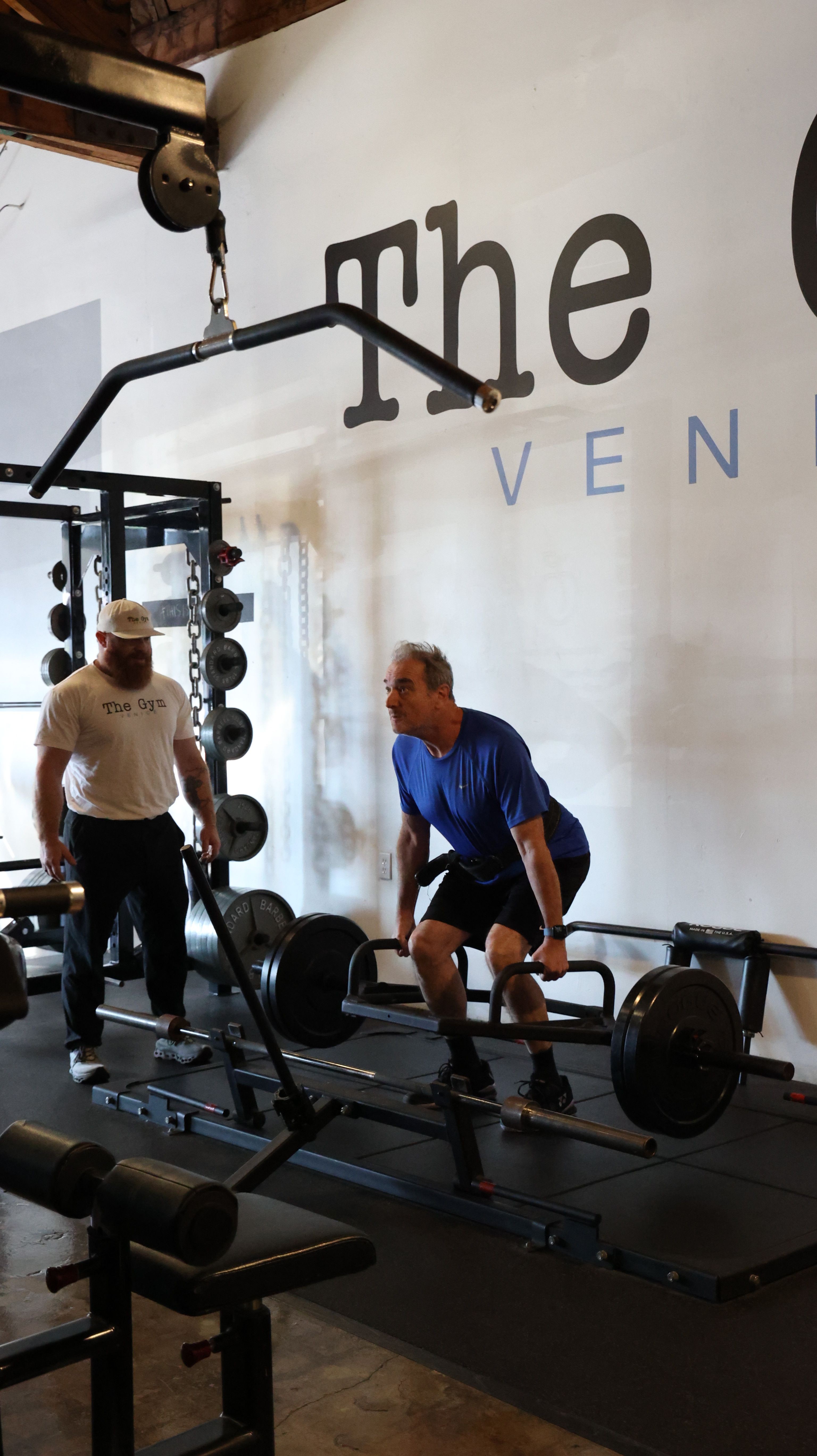 Two people in a gym near weightlifting equipment, one standing and one sitting, with a white wall and text in the background.