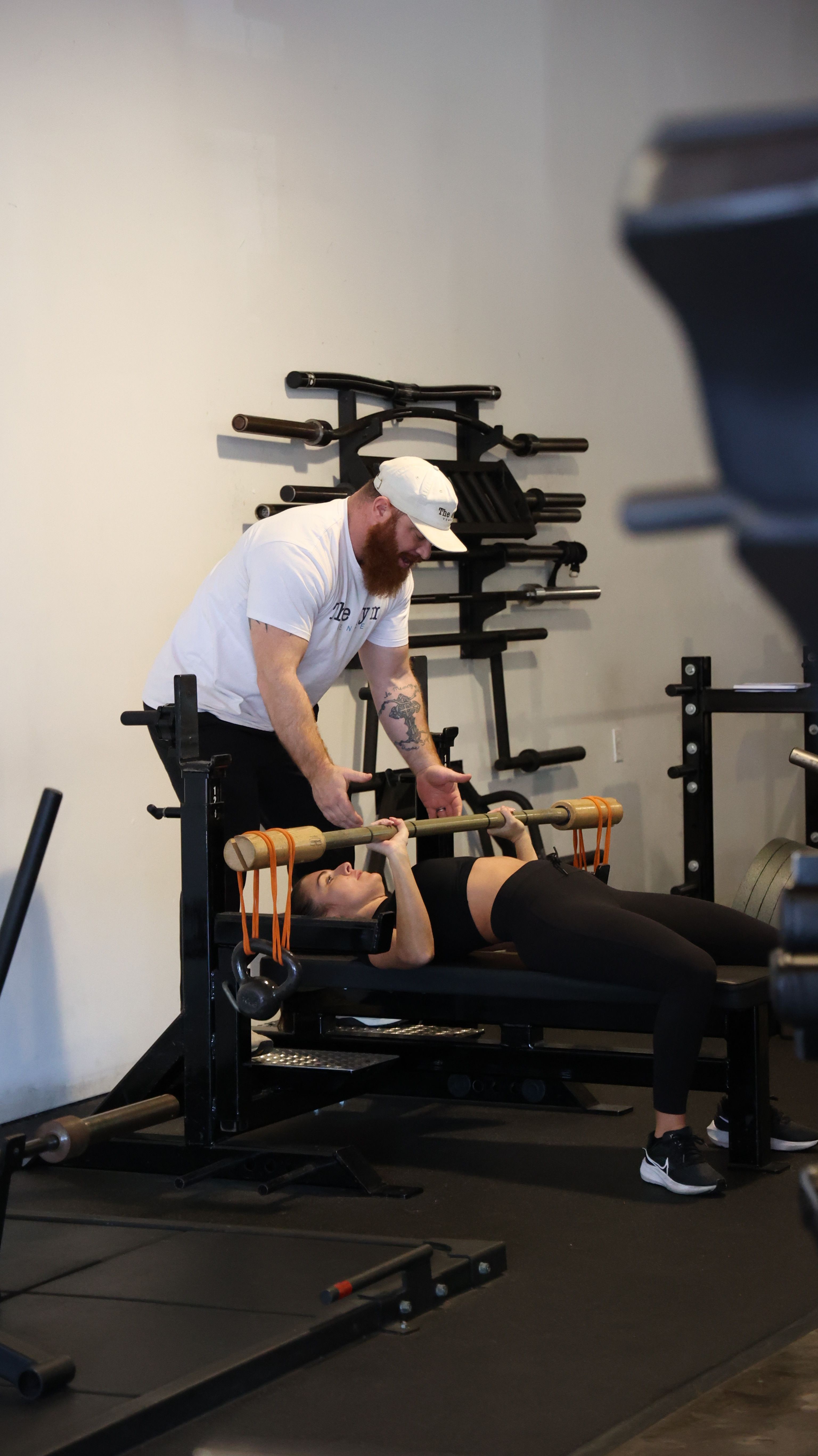 Person lying on a bench press with a trainer assisting in a gym setting.