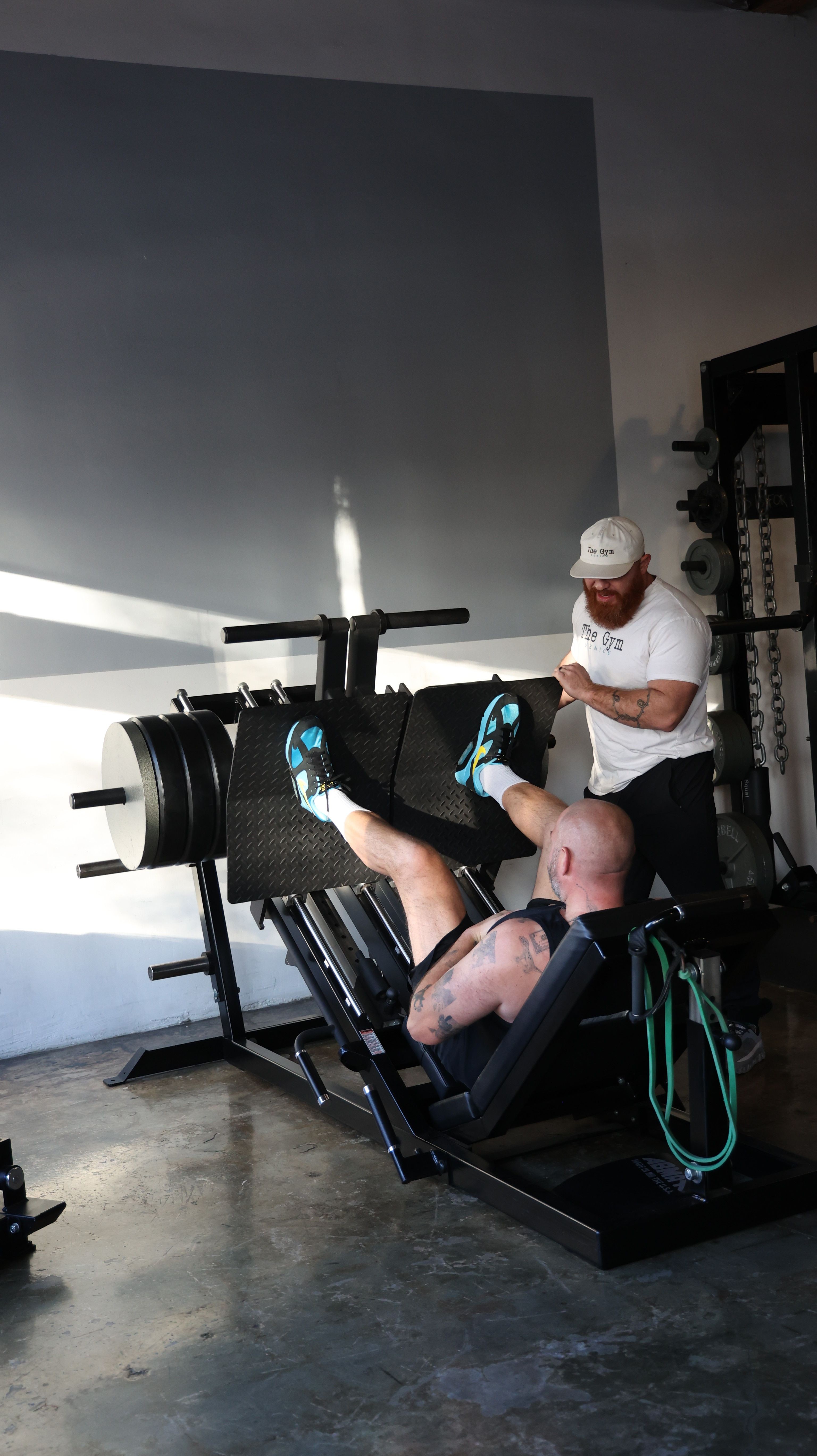 A man resting on a workout bench in a gym, with a woman standing nearby, gym equipment in background.