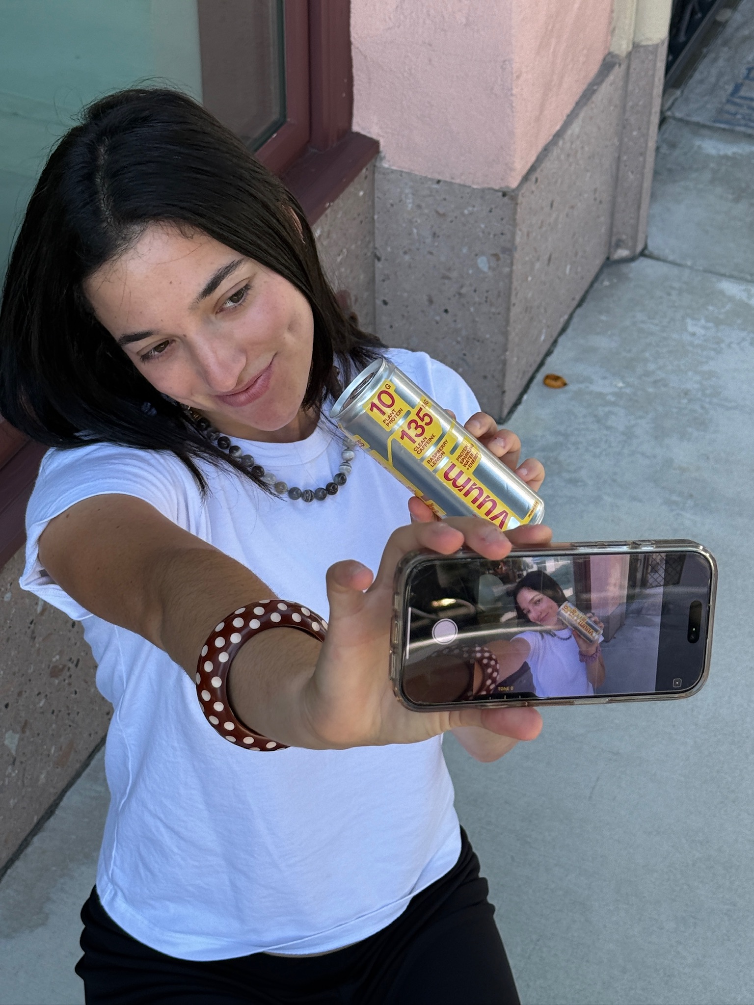Woman with dark hair taking a selfie with a smartphone outdoors, wearing a white shirt and a beaded bracelet.