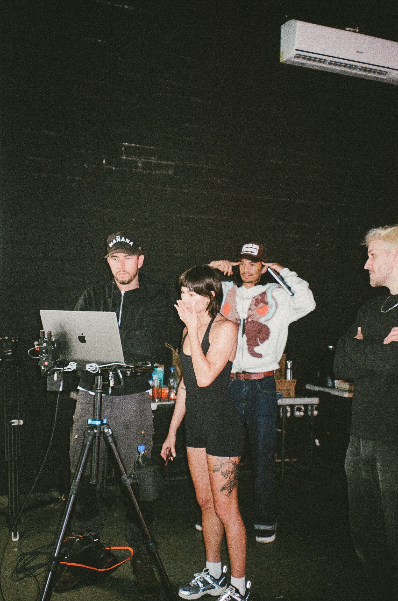 Four people standing in a dark room with a black wall, one person operating a camera, others watching, air conditioner above.