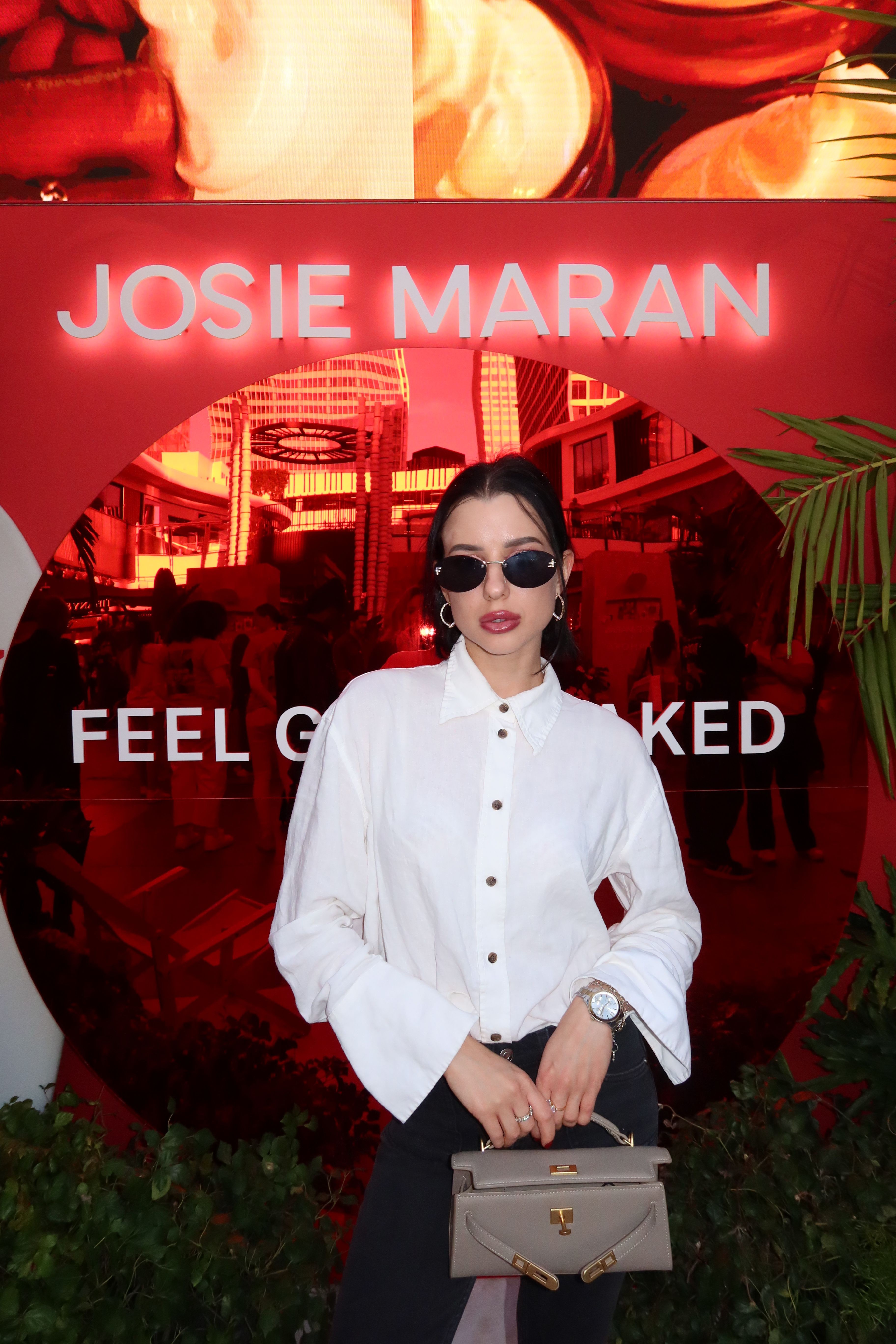 Woman in white shirt holding a gray handbag, standing in front of a red-lit background with palm leaves, text, and a building.