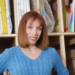Woman with shoulder-length red hair smiling, wearing a blue sweater, standing in front of shelves with fabric and supplies.