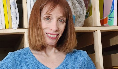 Woman with shoulder-length red hair smiling, wearing a blue sweater, standing in front of shelves with fabric and supplies.