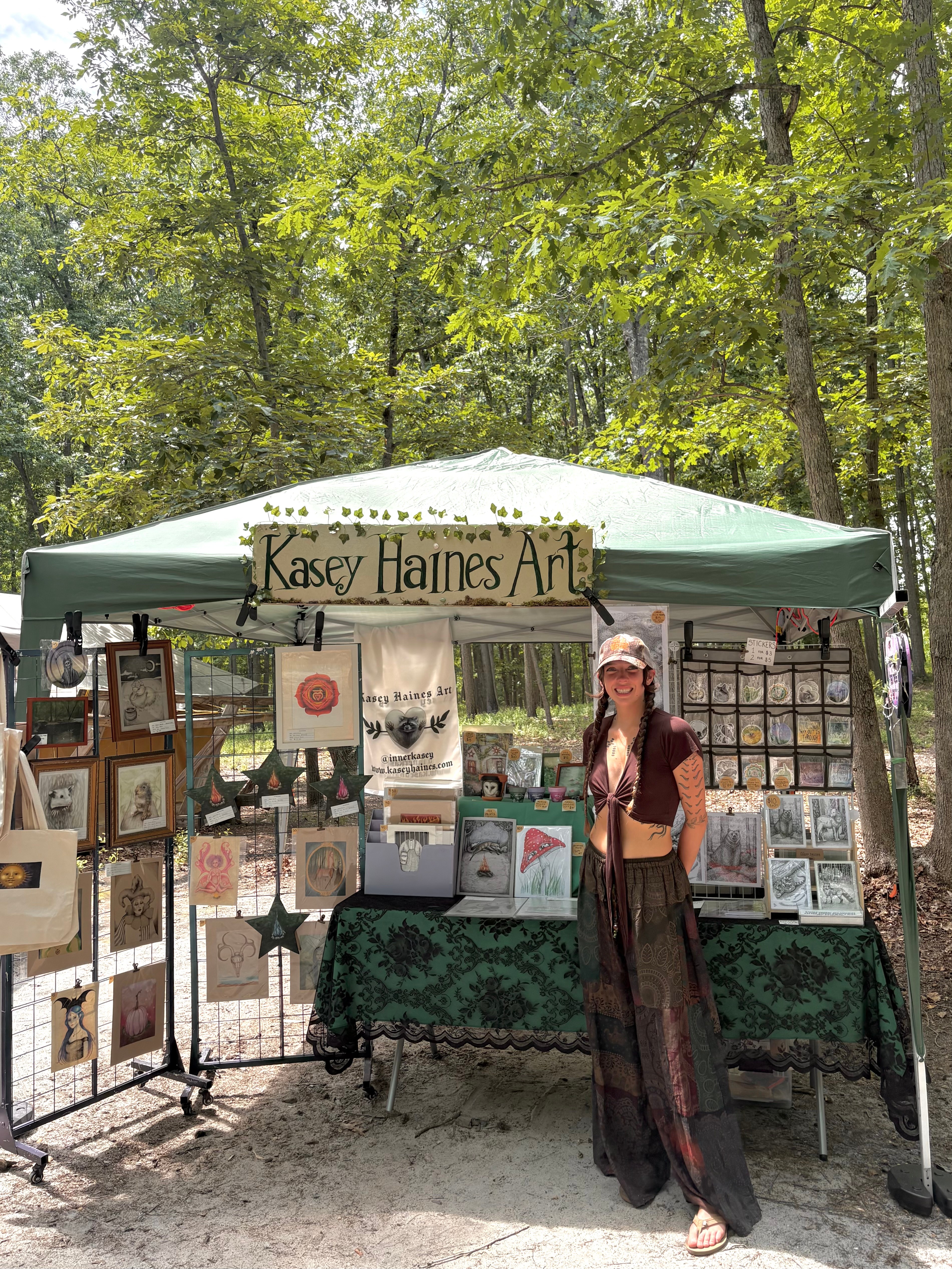 Woman standing in front of an art booth with framed artwork, trees in the background, and a sign reading