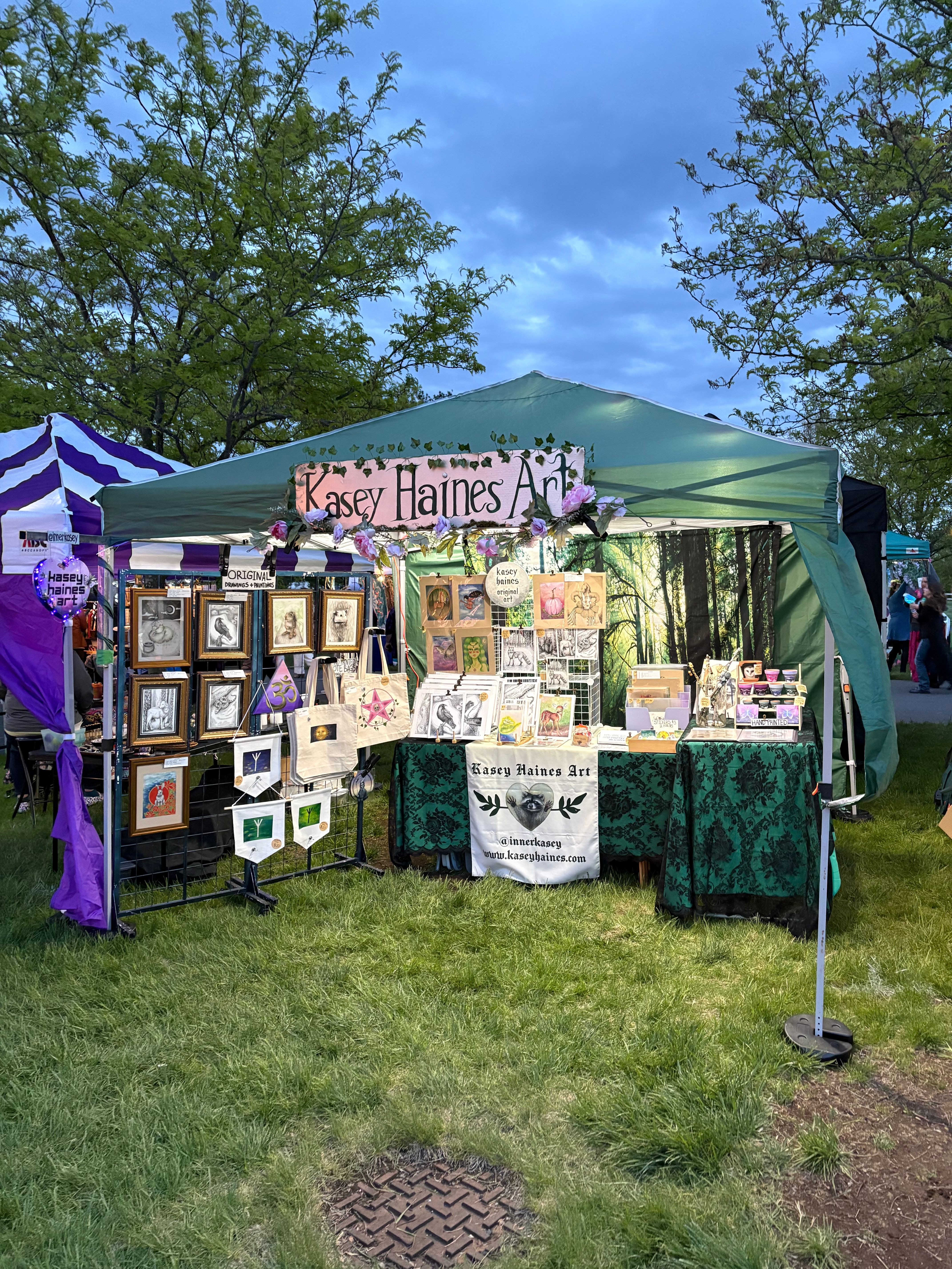 Outdoor art display with framed pictures and artwork under a green canopy, surrounded by trees and grass.
