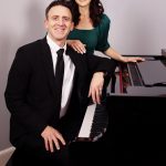 A man in a black suit and a woman in a green top sit at a piano, smiling, in a room with a staircase.