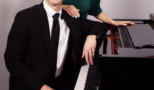 A man in a black suit and a woman in a green top sit at a piano, smiling, in a room with a staircase.