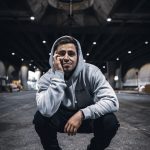 Young man crouching outdoors at night, wearing a hoodie and glasses, with a bridge structure overhead and city lights in background.