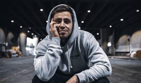 Young man crouching outdoors at night, wearing a hoodie and glasses, with a bridge structure overhead and city lights in background.