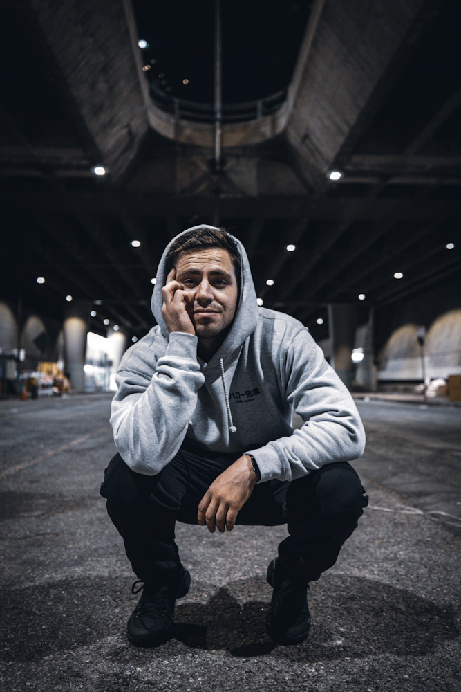 Young man crouching outdoors at night, wearing a hoodie and glasses, with a bridge structure overhead and city lights in background.