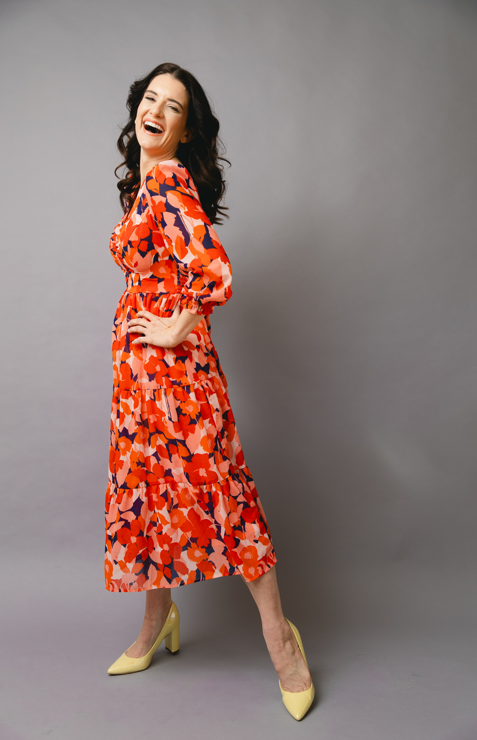 Woman in a colorful patterned dress laughing, standing with one hand on her hip, against a gray background.