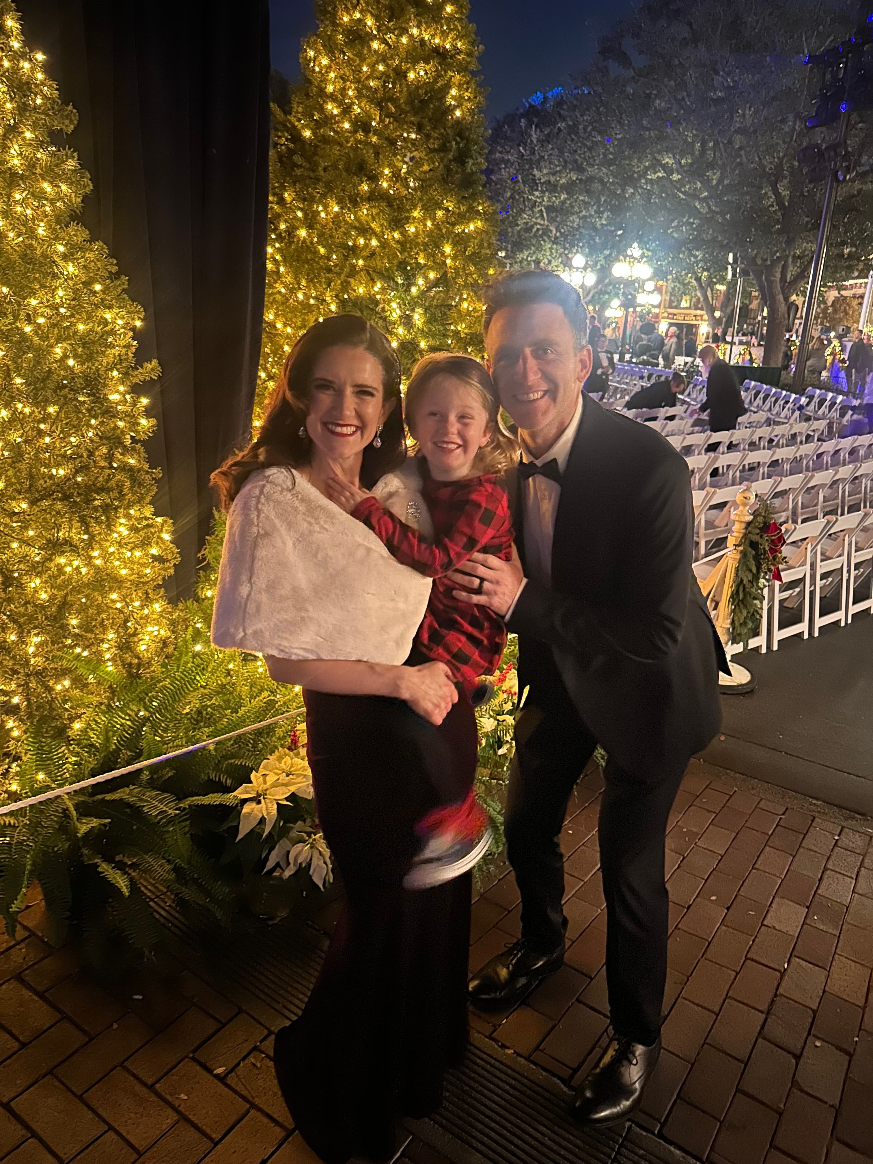 Three people standing outdoors at night near a decorated Christmas tree with lights, smiling and posing together.
