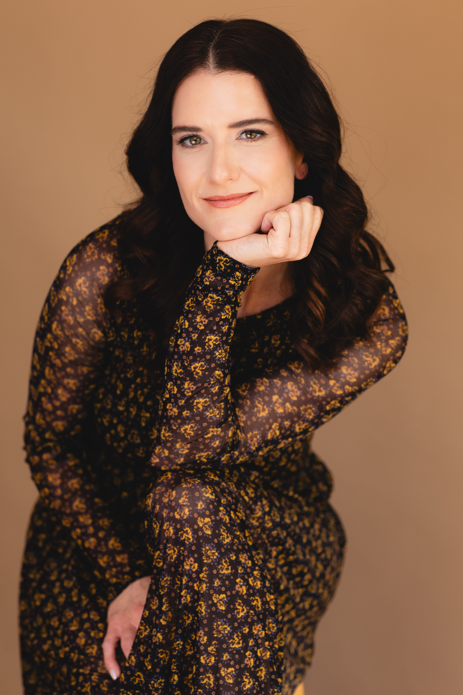Woman with long dark hair, smiling, resting her chin on her hand, wearing a floral patterned dress, beige background.