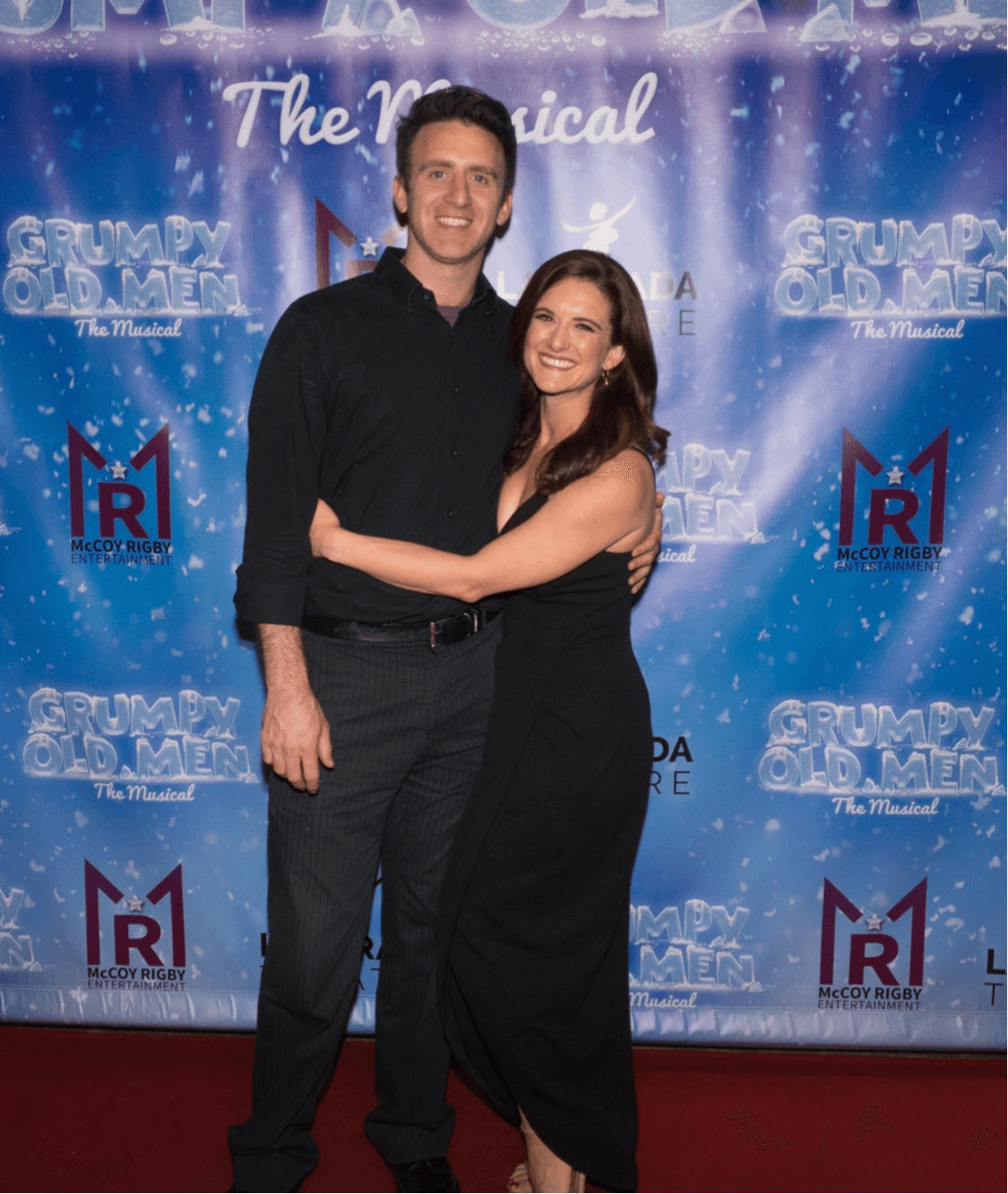 A man and woman stand together smiling, with a blue backdrop and event logos behind them.