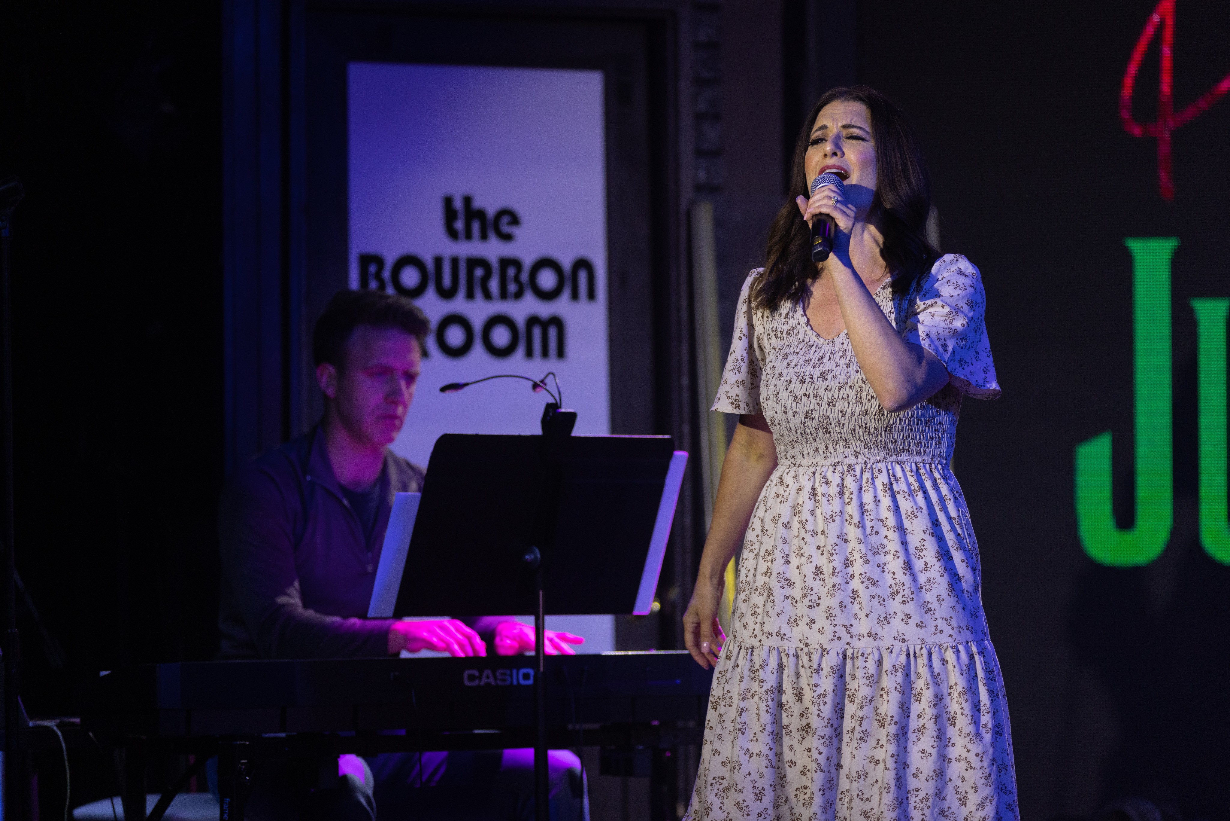 A woman sings into a microphone on stage, with a man playing keyboard beside her, in a dimly lit venue.