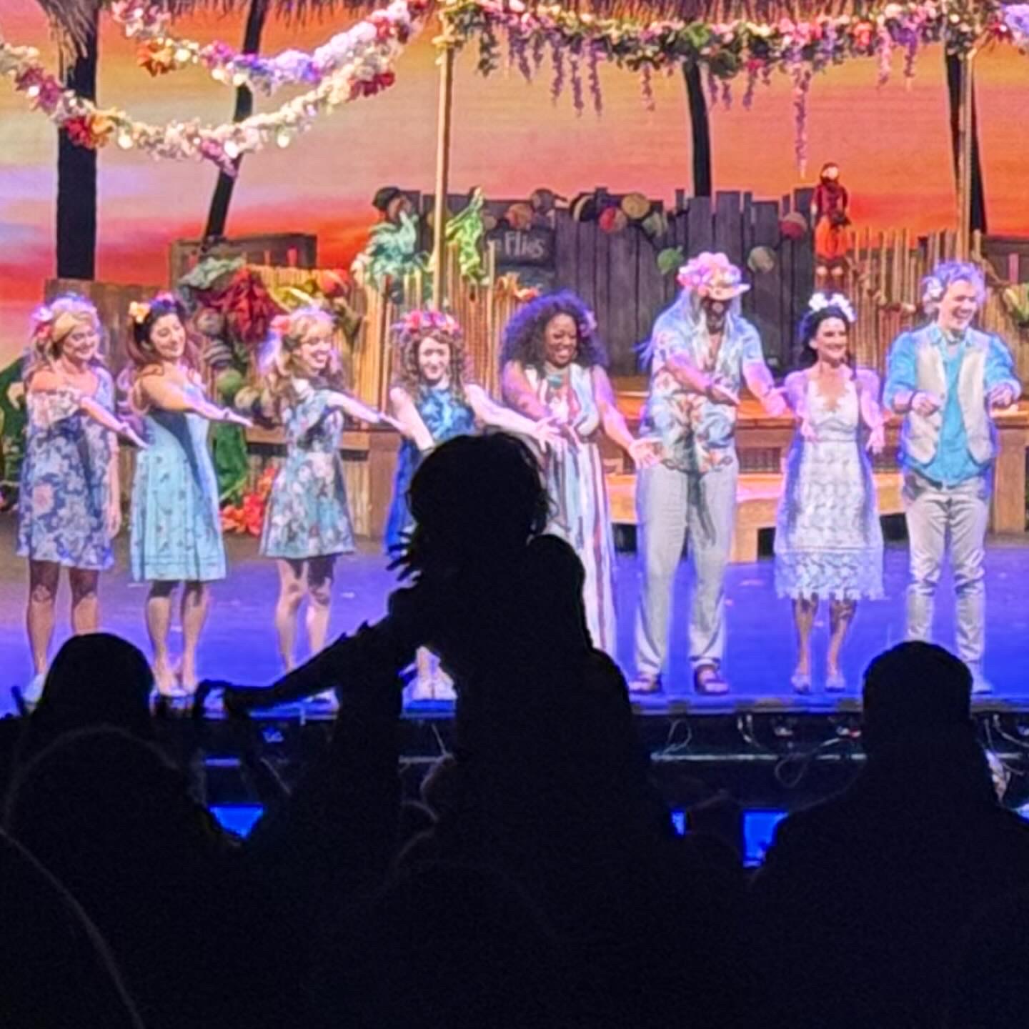Group of children performing on stage with colorful decorations and a sunset backdrop, holding hands and smiling.