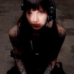 Young woman with dark hair and tattoos wearing headphones, looking up at the camera, dark background.