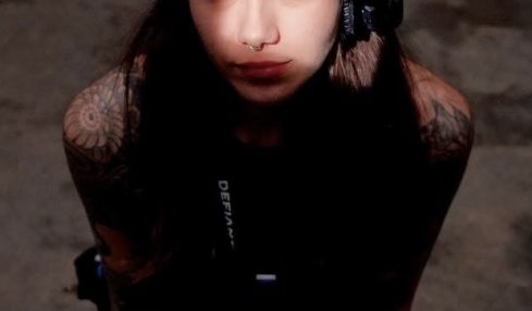 Young woman with dark hair and tattoos wearing headphones, looking up at the camera, dark background.