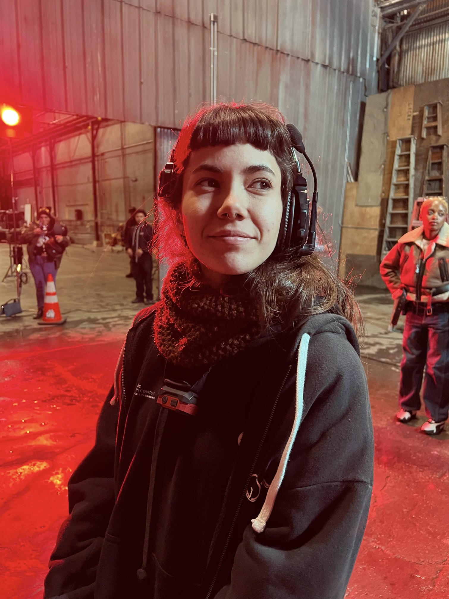 Young woman with headphones and scarf, smiling, in an industrial setting with red lighting and people in the background.