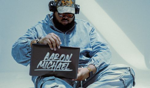 Person sitting cross-legged on the floor, wearing a helmet with headphones, holding a sign that says 'Aaron Michael', with red circular objects nearby.
