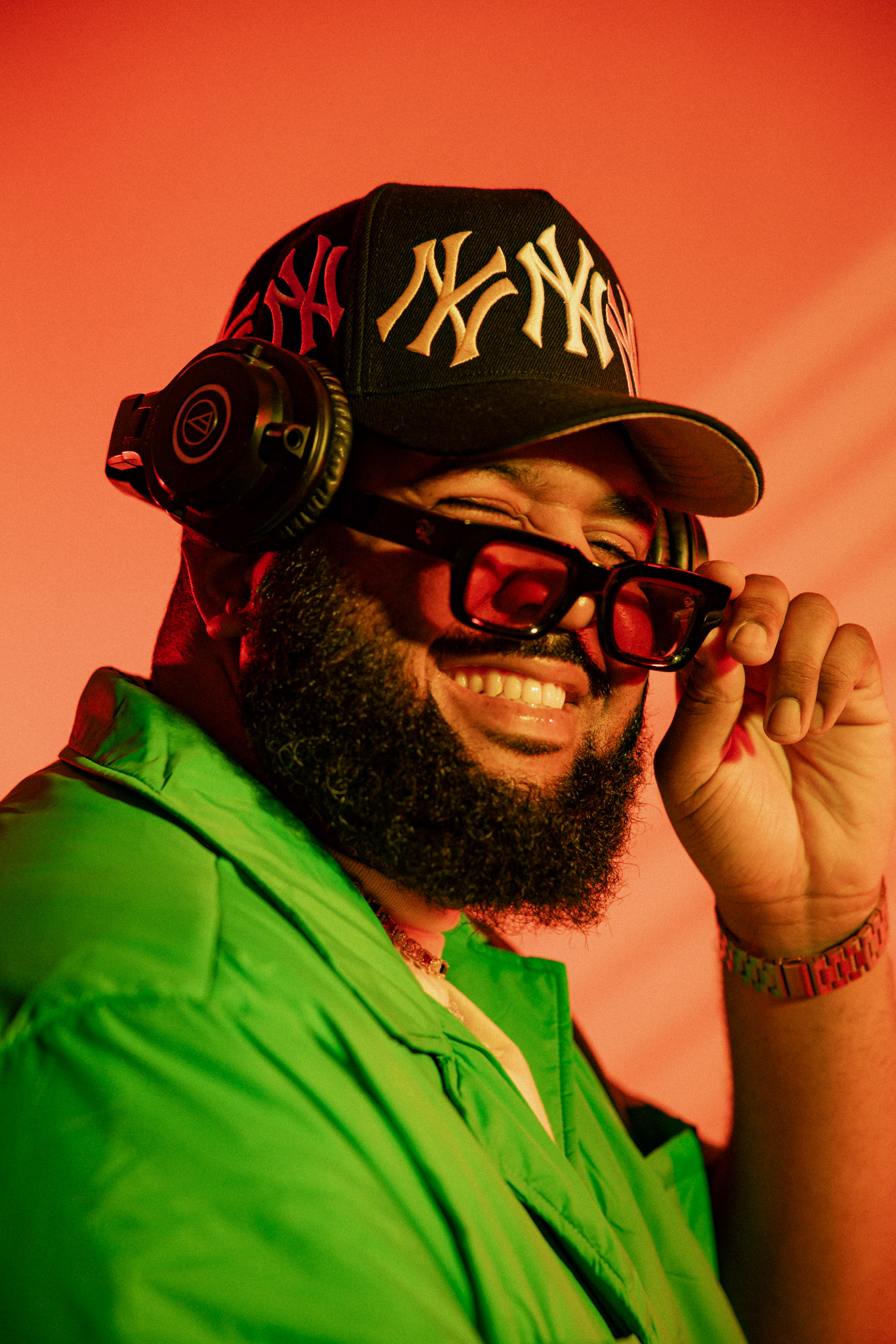 Person wearing a black New York Yankees cap, red glasses, green jacket, smiling, holding glasses, with headphones, orange background.