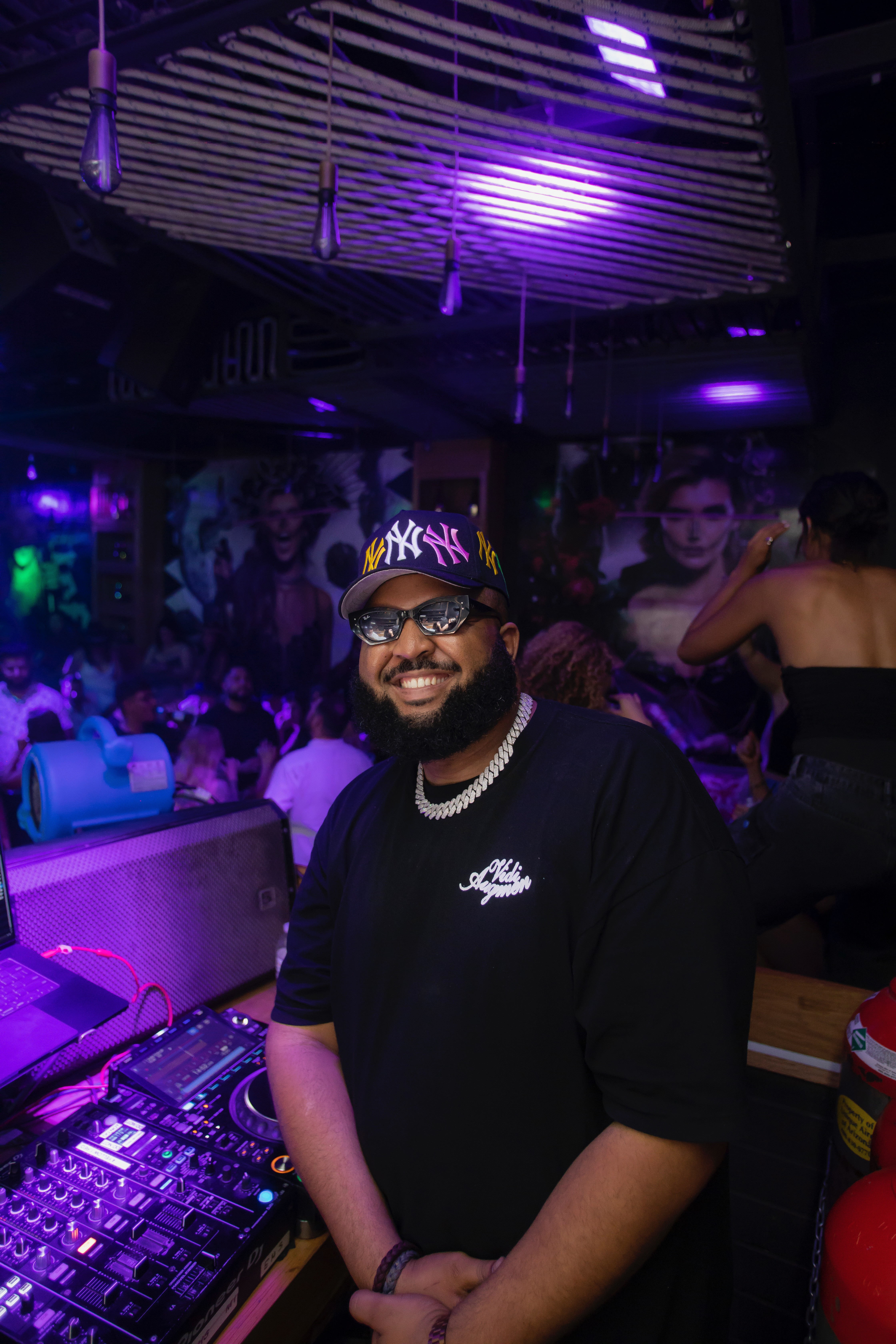 Man with glasses and a cap smiling in a nightclub with purple lighting and people dancing in the background.