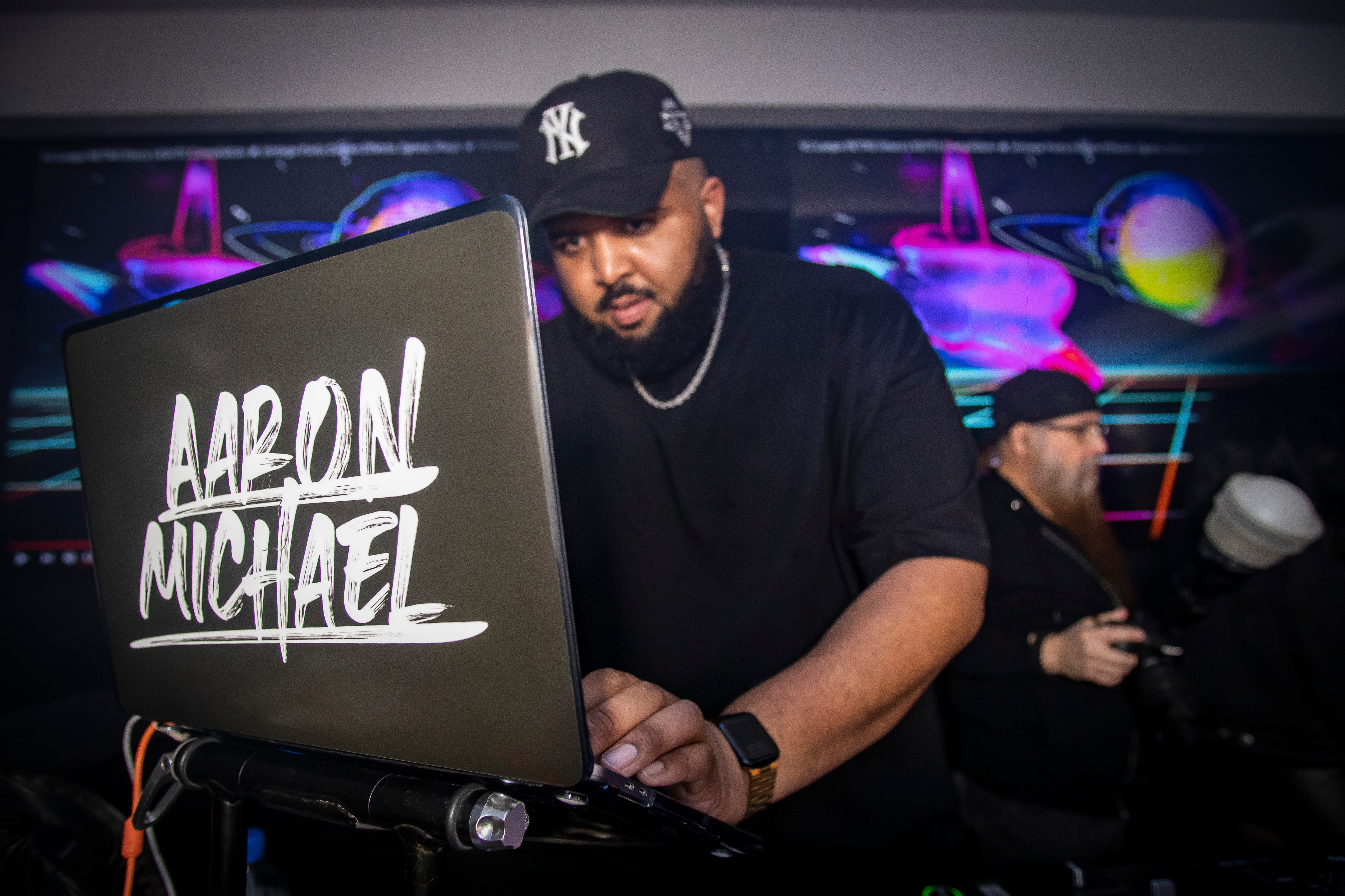 Man wearing a black cap and shirt working on a laptop with a sign reading 'Aaron Michael' in a club or event setting.
