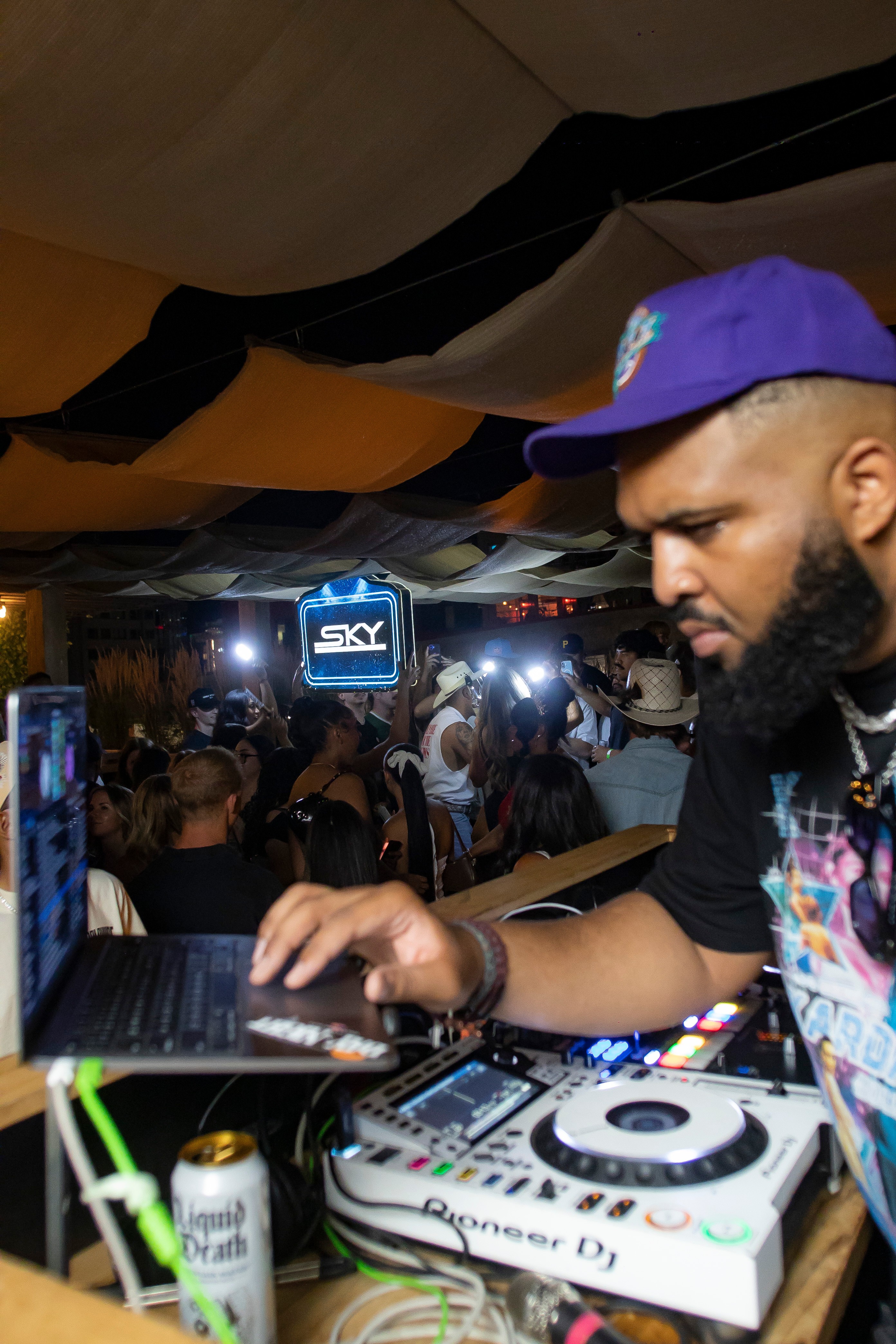 DJ with a beard wearing a blue cap and colorful shirt, operating equipment at a crowded indoor event with a screen displaying 'SKY'.