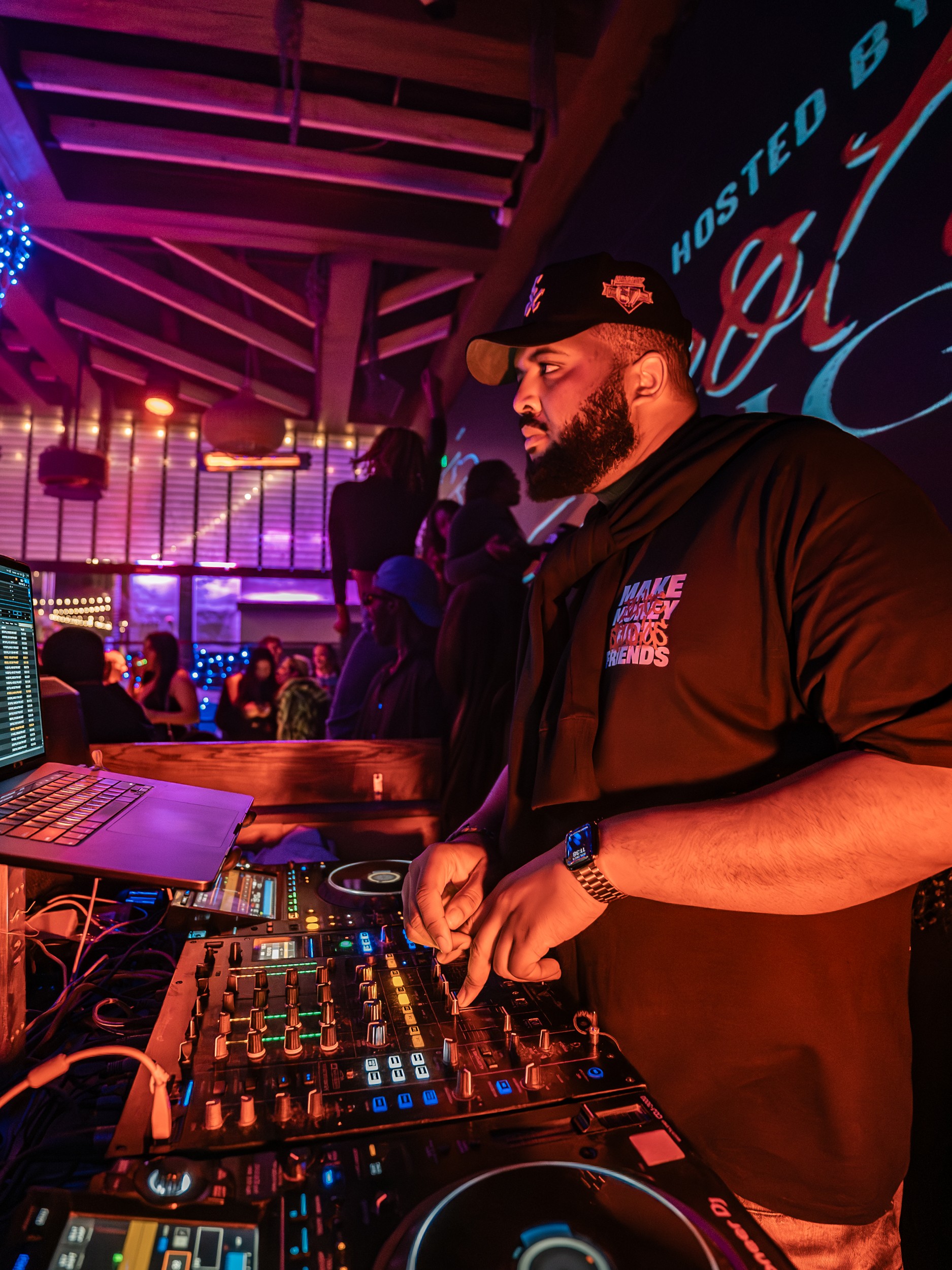 Bearded DJ wearing a black cap and hoodie, operating a DJ mixer in a lively indoor venue with colorful lighting.