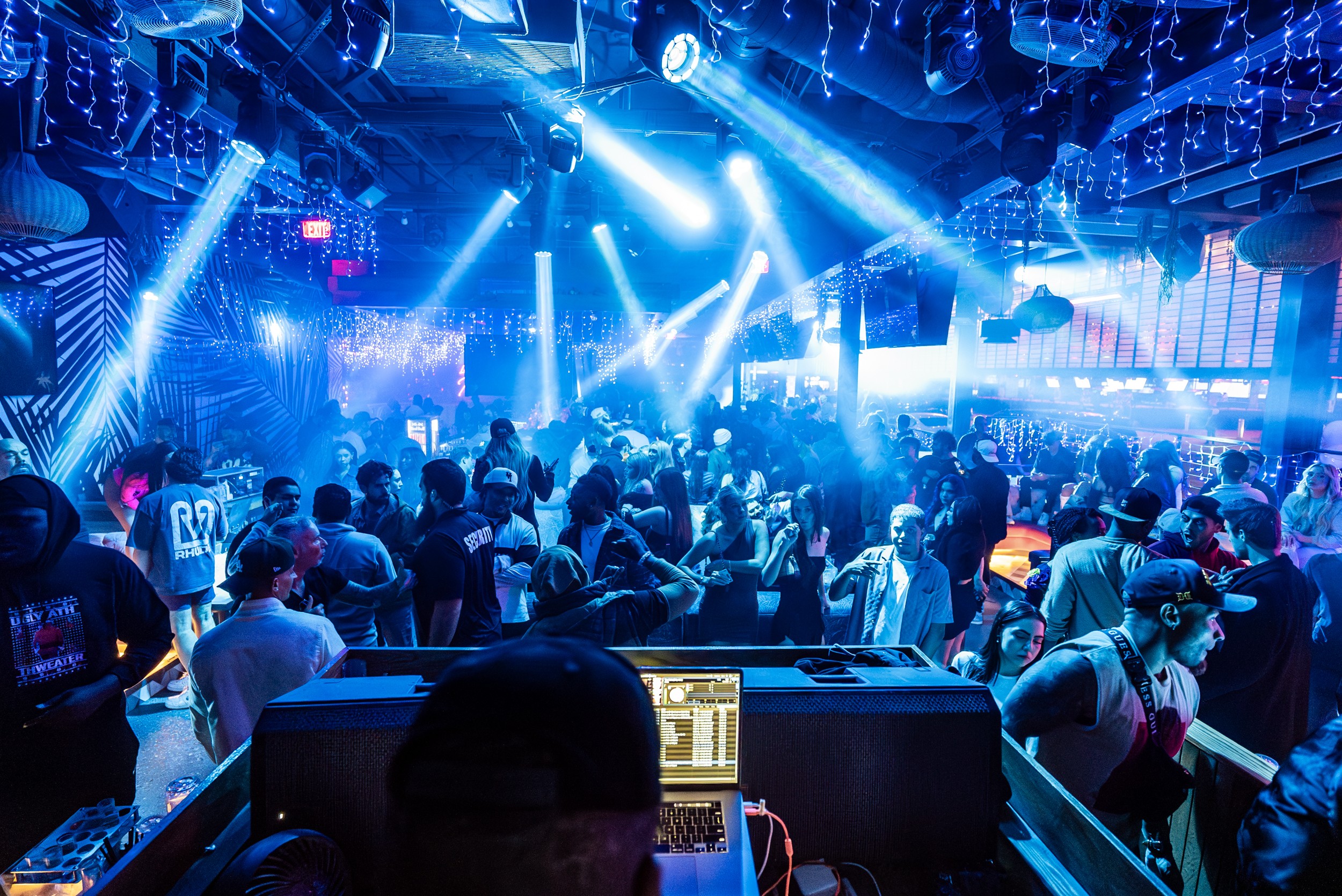 Crowd dancing in a nightclub with bright blue and white lights, DJ booth in foreground, people enjoying music.