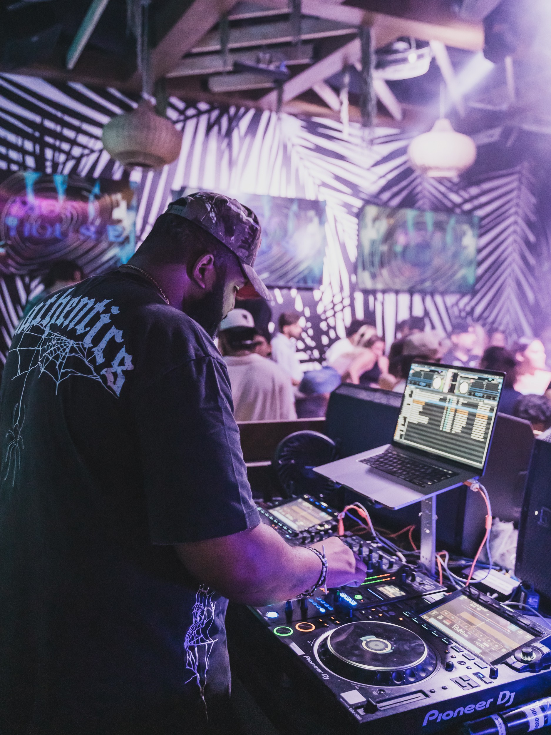 DJ working with equipment in a colorful, lively club with people dancing and vibrant lighting.